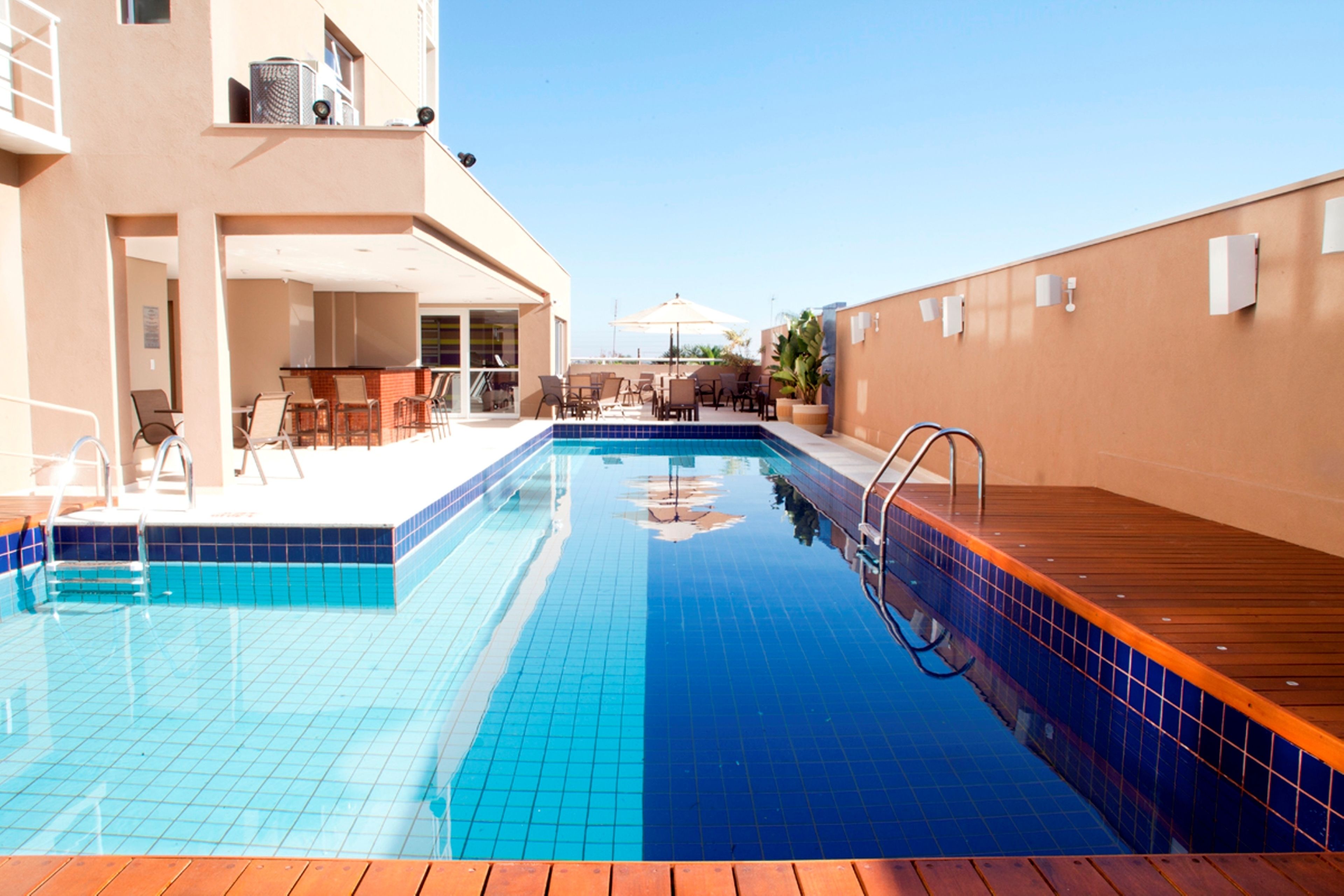 Une piscine aux carreaux bleus, une terrasse en bois et un espace extérieur aménagé avec des chaises et des parasols.