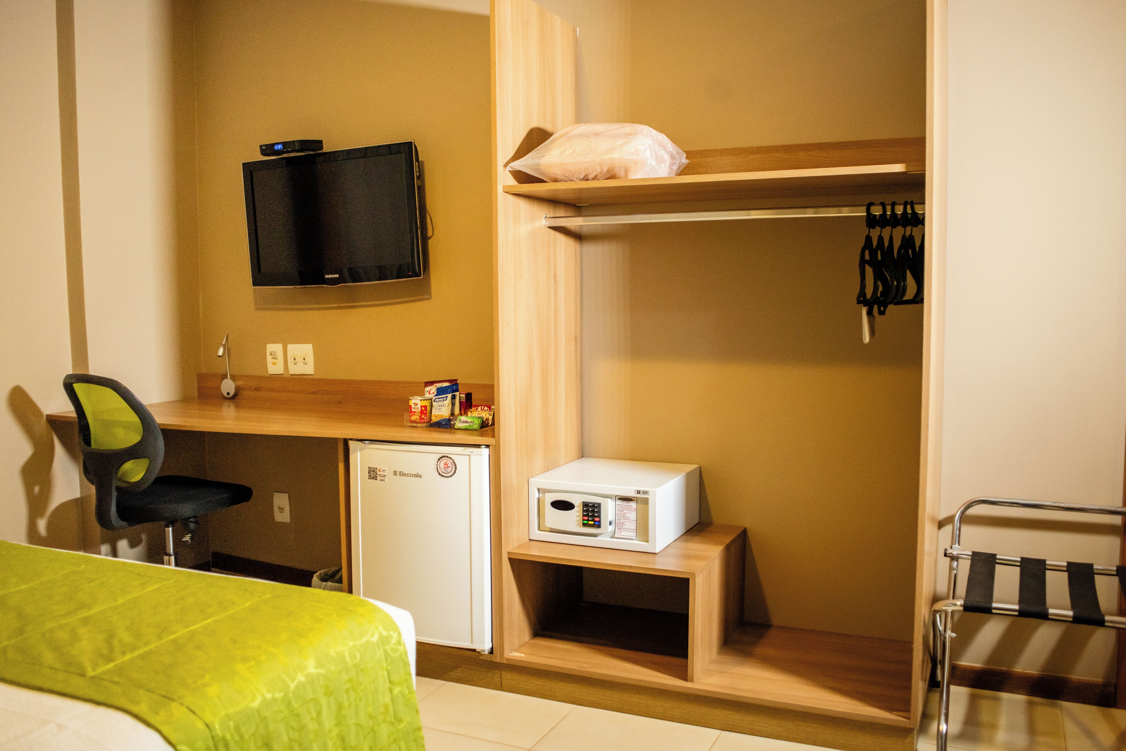 A compact hotel room with a bed, a desk with a computer, a small fridge, a microwave, and a TV mounted on the wall.