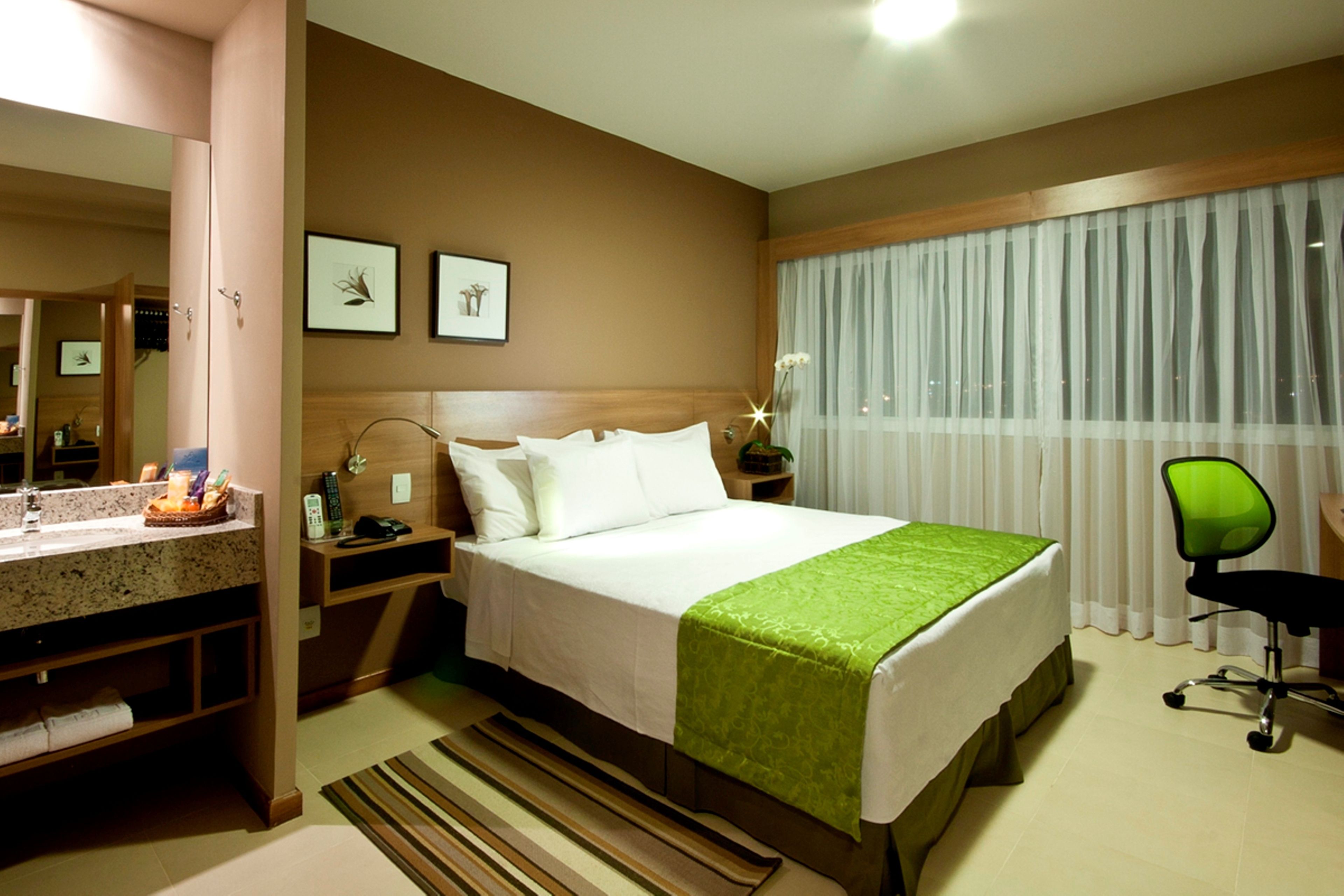 Une chambre d’hôtel avec un lit, un bureau, une chaise, un miroir et une fenêtre avec des rideaux.