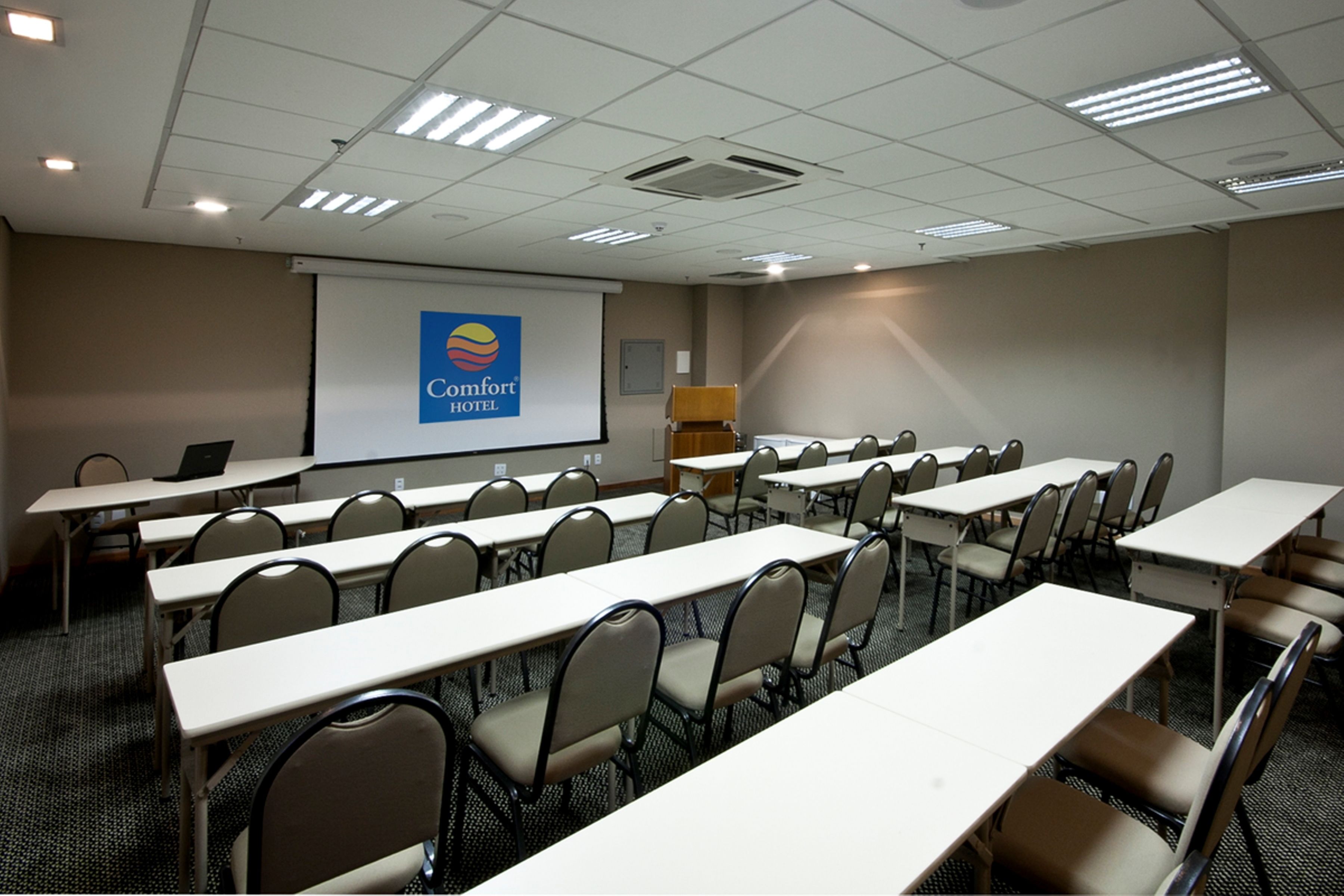 Un salón de conferencias con filas de mesas y sillas, una pantalla de proyección con el logotipo de Comfort Hotel y una pizarra.