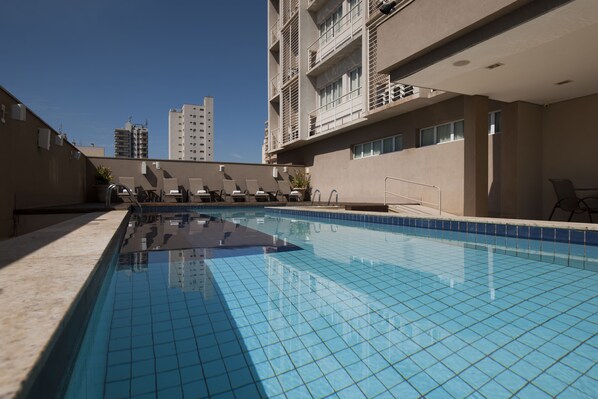 Una piscina con baldosas azules cristalinas, rodeada de sillas de descanso y un edificio con balcones.