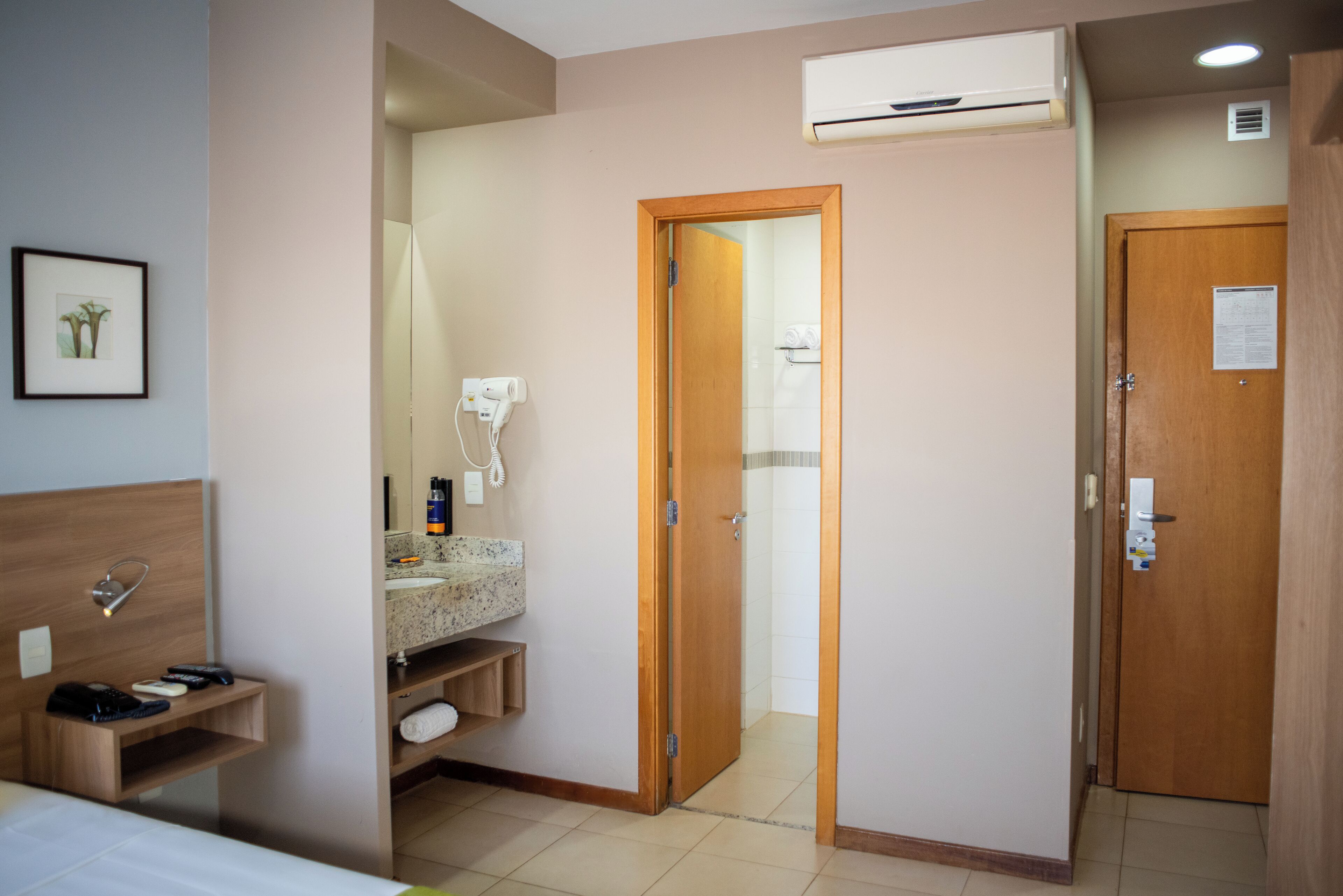 A hotel room with a wooden headboard, a built-in shelf, a bathroom with a sink and mirror, and a door leading to another room.