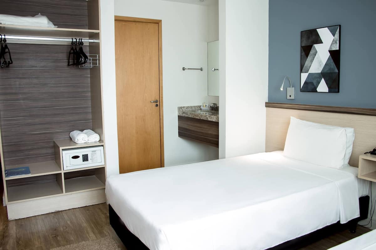 A hotel room with a bed, a wardrobe, a bedside table, a mirror, and a wall art piece.
