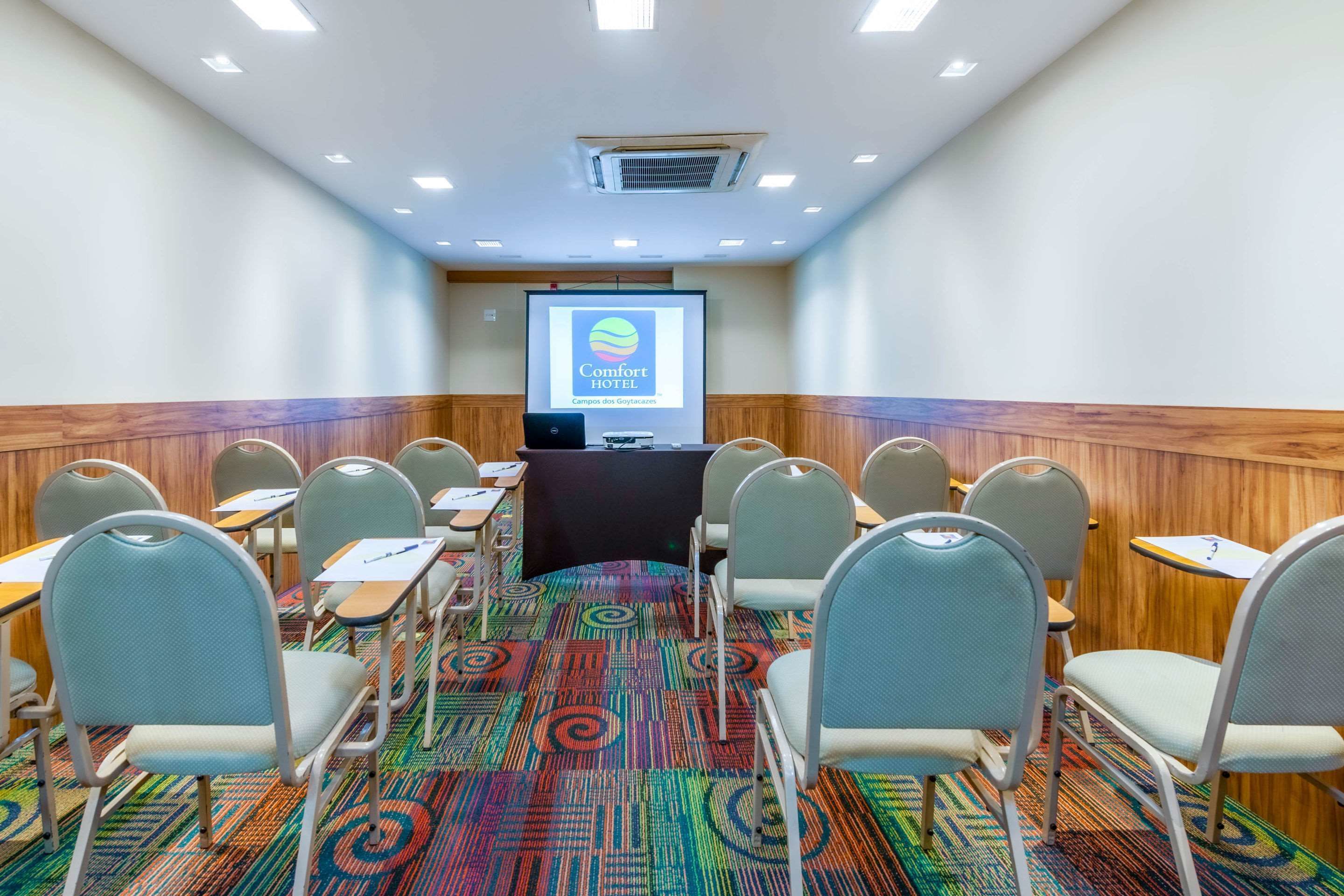 A conference room with a large screen displaying the Comfort Hotel logo, a carpeted floor, and rows of chairs.