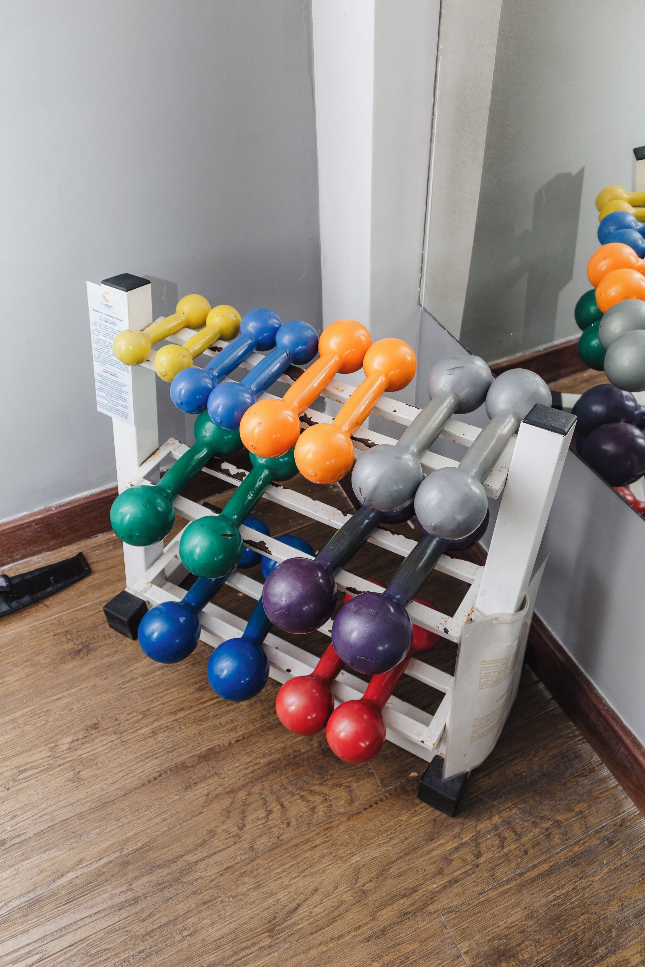 A rack of colorful dumbbells in a room with wooden flooring and a white wall.