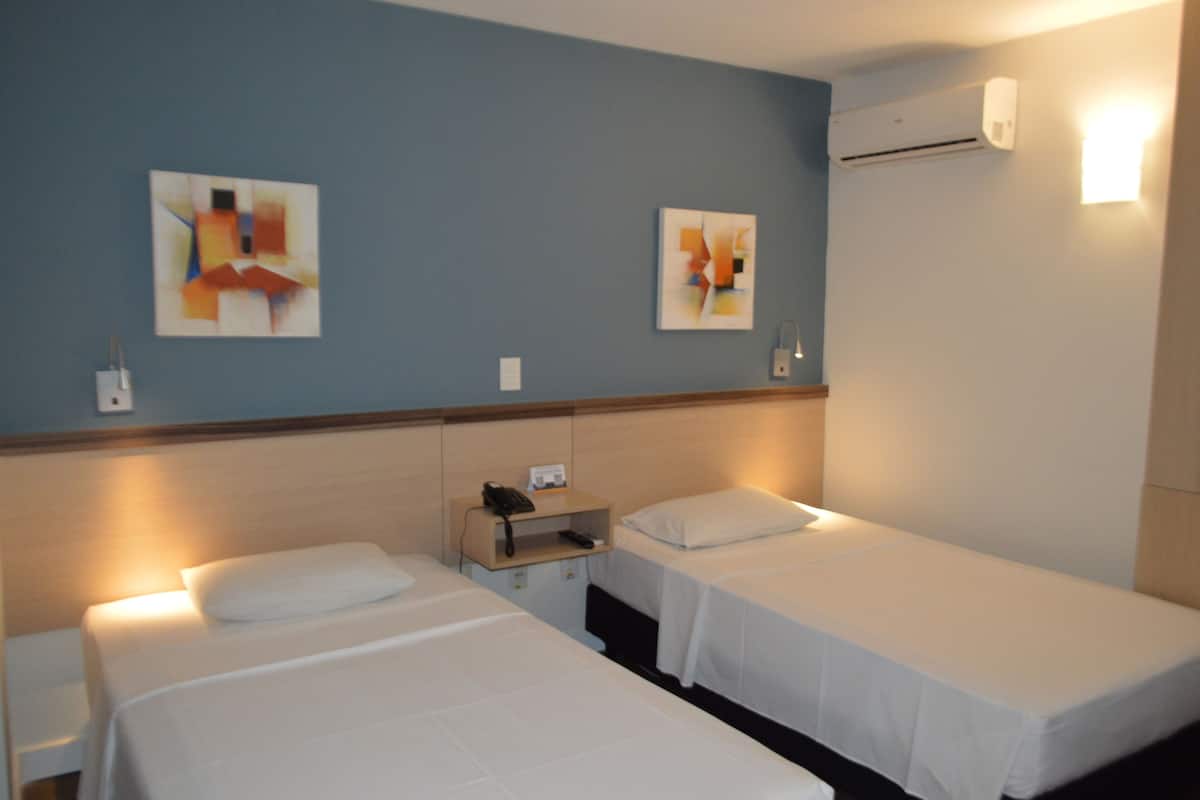 A hotel room with two single beds, a bedside table with a phone and lamp, and abstract art on the walls.