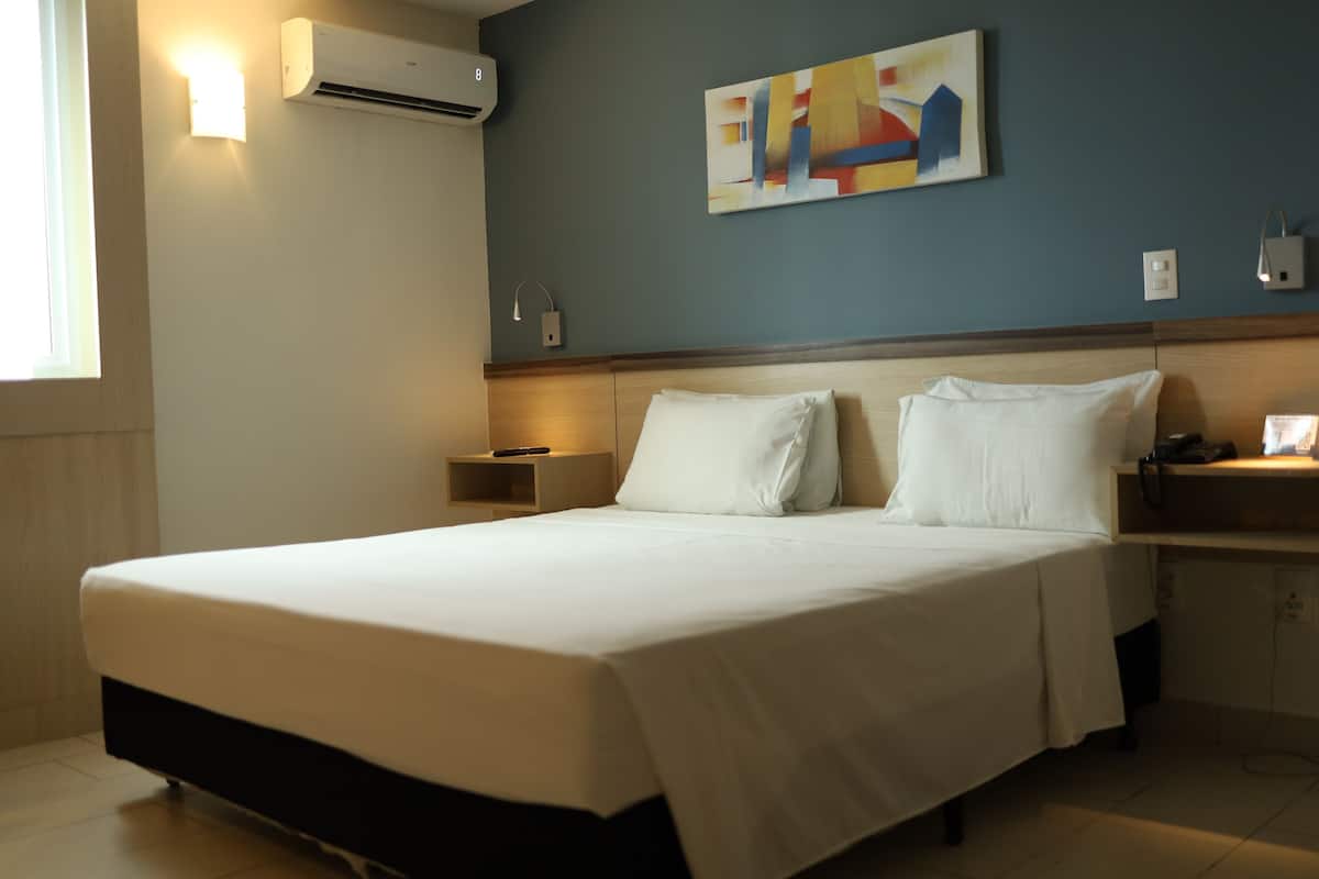A hotel room with a large bed, a nightstand, a wall-mounted air conditioner, a painting, and a bedside lamp.