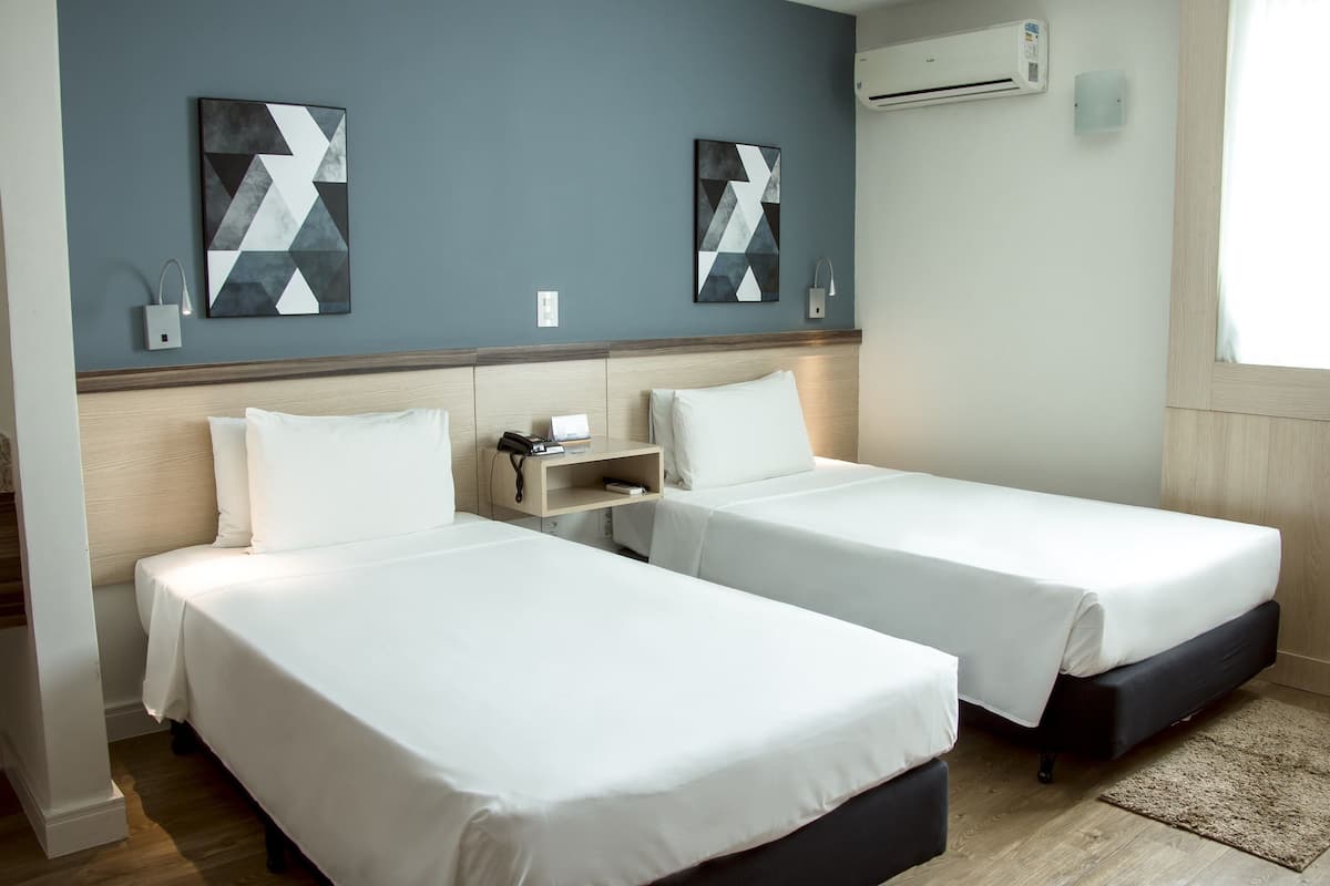 A hotel room with two beds, a nightstand, an air conditioning unit, and two abstract paintings on the wall.
