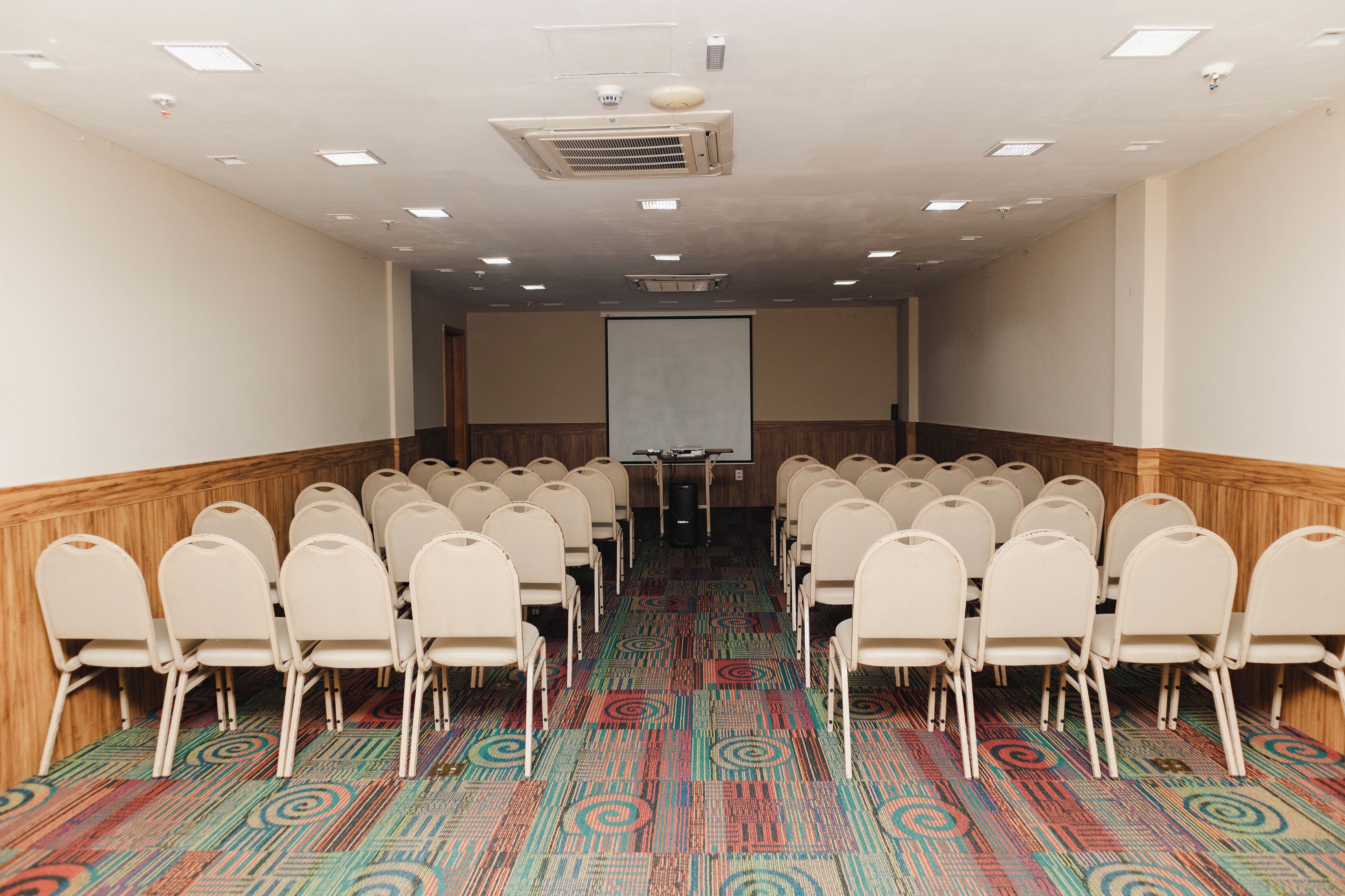 Uma sala de conferências com fileiras de cadeiras, uma tela de projetor e piso coberto de carpete.