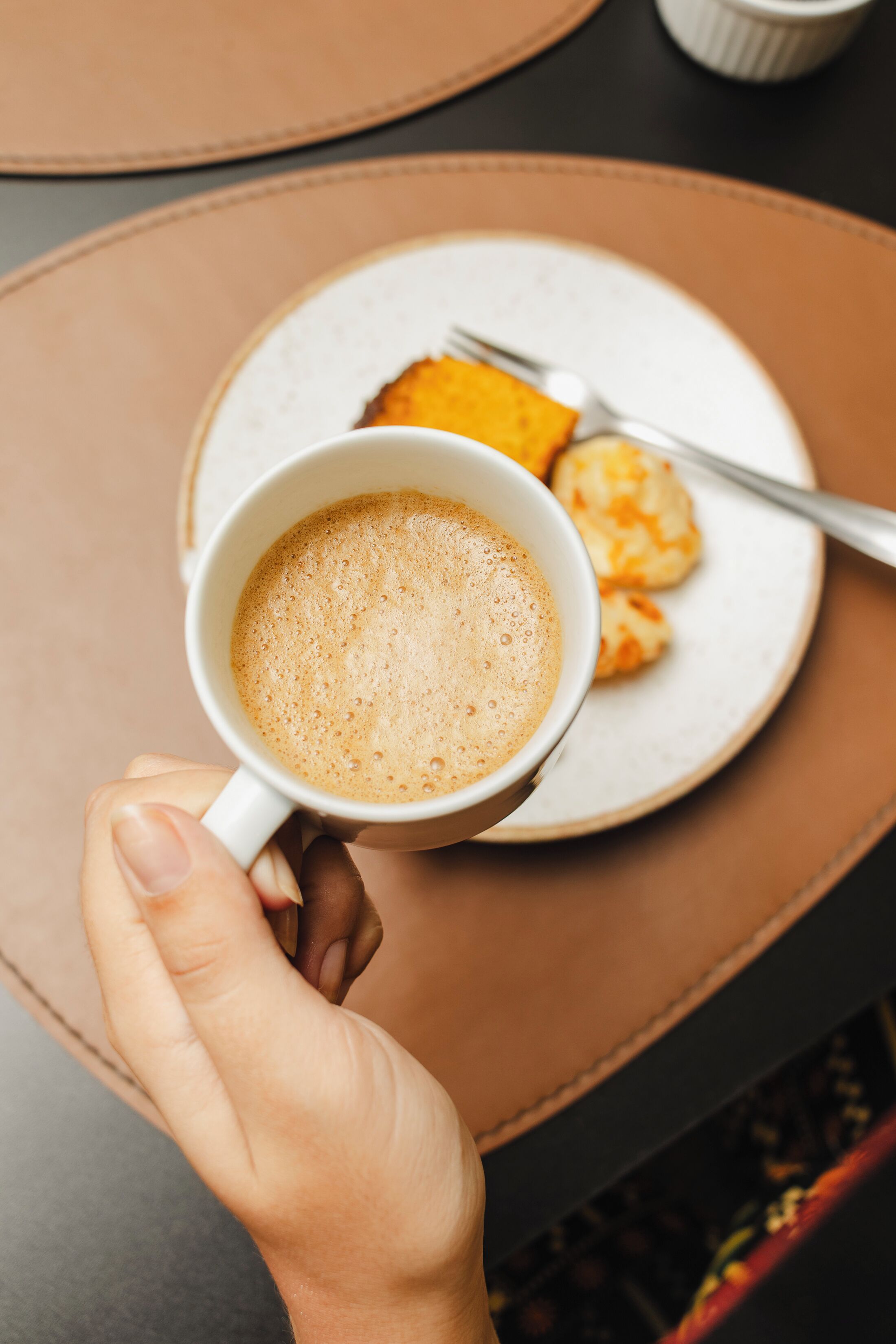 A person holding a cup of coffee with a plate of food in the background.