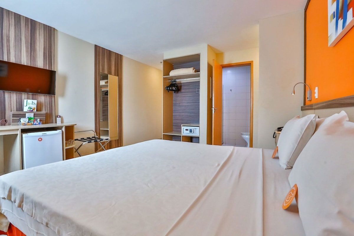 Habitación de hotel con cama, escritorio, mini refrigerador, armario y zona de ducha.