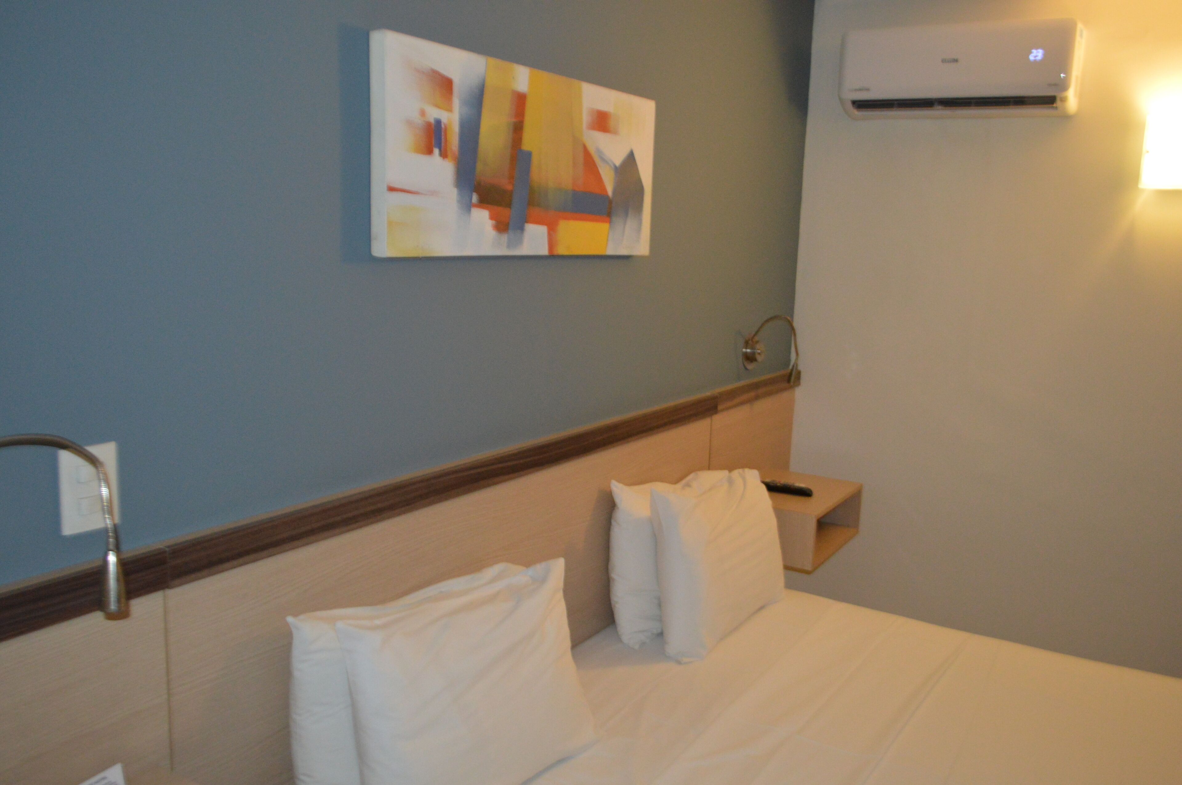 Een hotelkamer met een bed, kussens, een wandairconditioner, een schilderij en een nachtkastje.