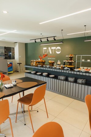 Un moderno comedor con una barra que exhibe diversas opciones de comida, un espacio con sillas y una pared con la palabra 'EATS' que indica una zona de comedor.