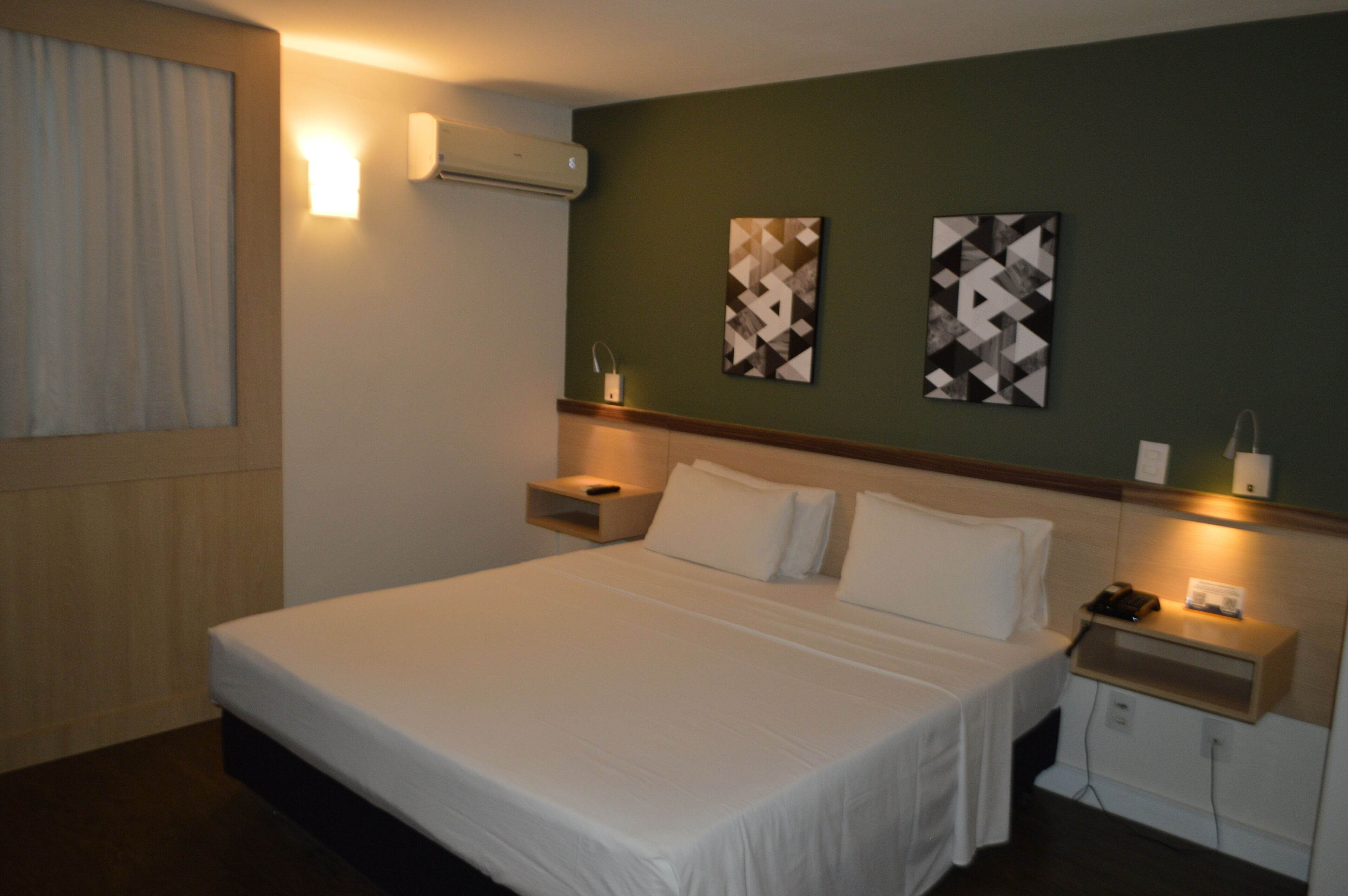 Une chambre d’hôtel avec un grand lit, des tables de chevet, un climatiseur fixé au mur et deux œuvres d’art abstraites accrochées au mur.