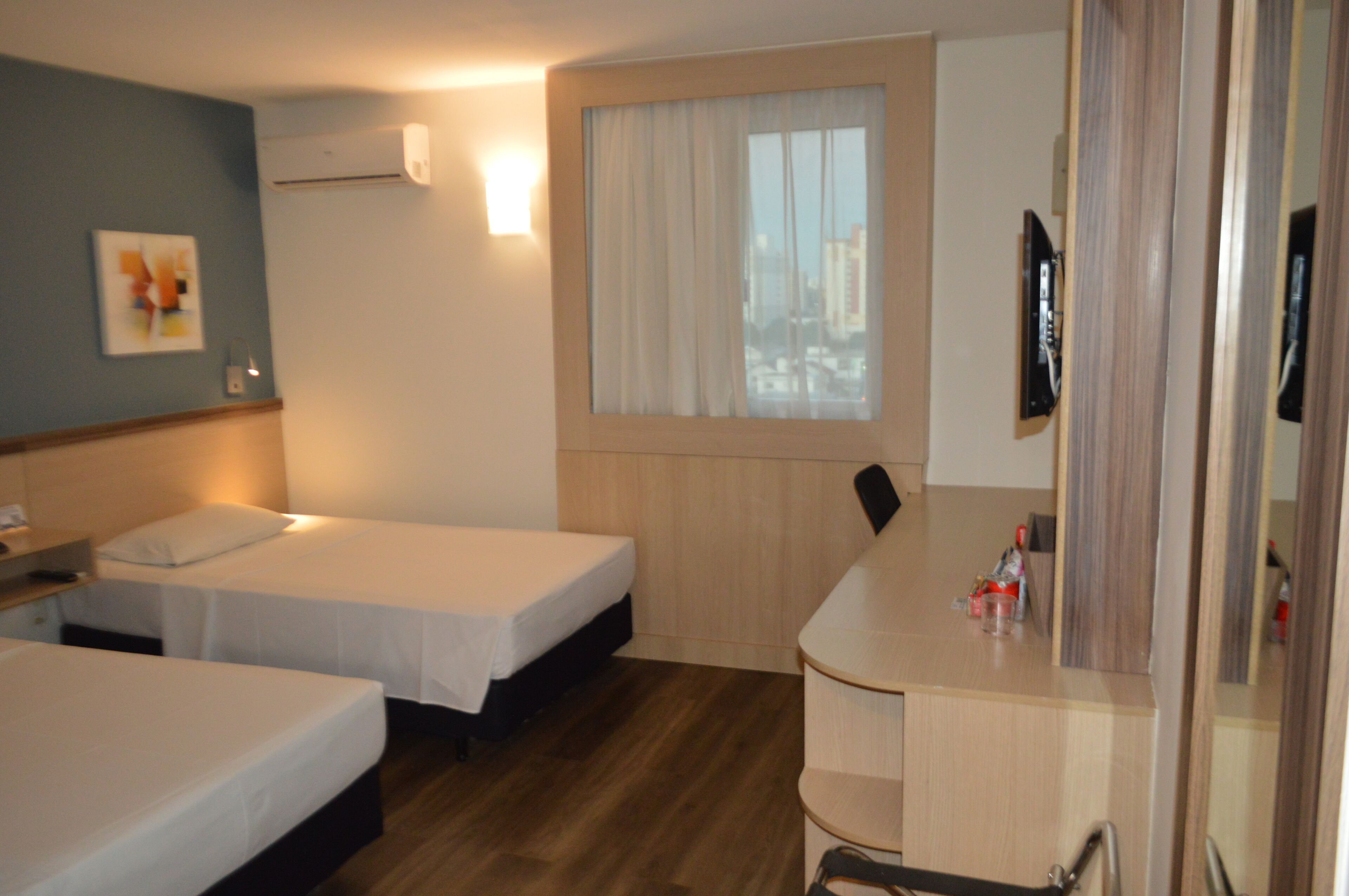 Ein Hotelzimmer mit zwei Betten, einem Schreibtisch mit Spiegel, einem Fenster mit Vorhängen und einer Klimaanlage an der Wand.