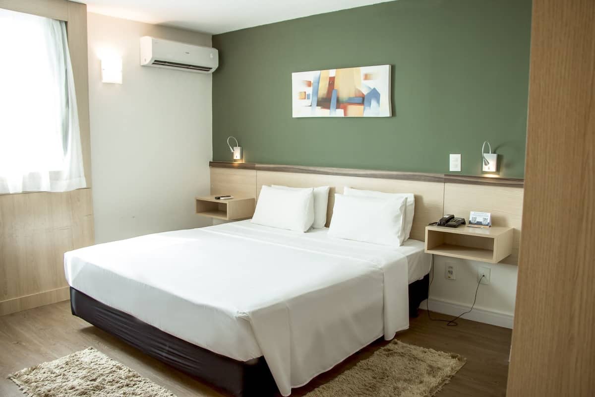 A hotel room with a large bed, bedside tables, a wall-mounted air conditioner, a painting, and a small shelf with a phone and a lamp.