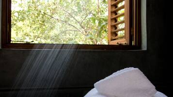 A window with wooden shutters, a view of green foliage outside, and two rolled towels on a ledge.