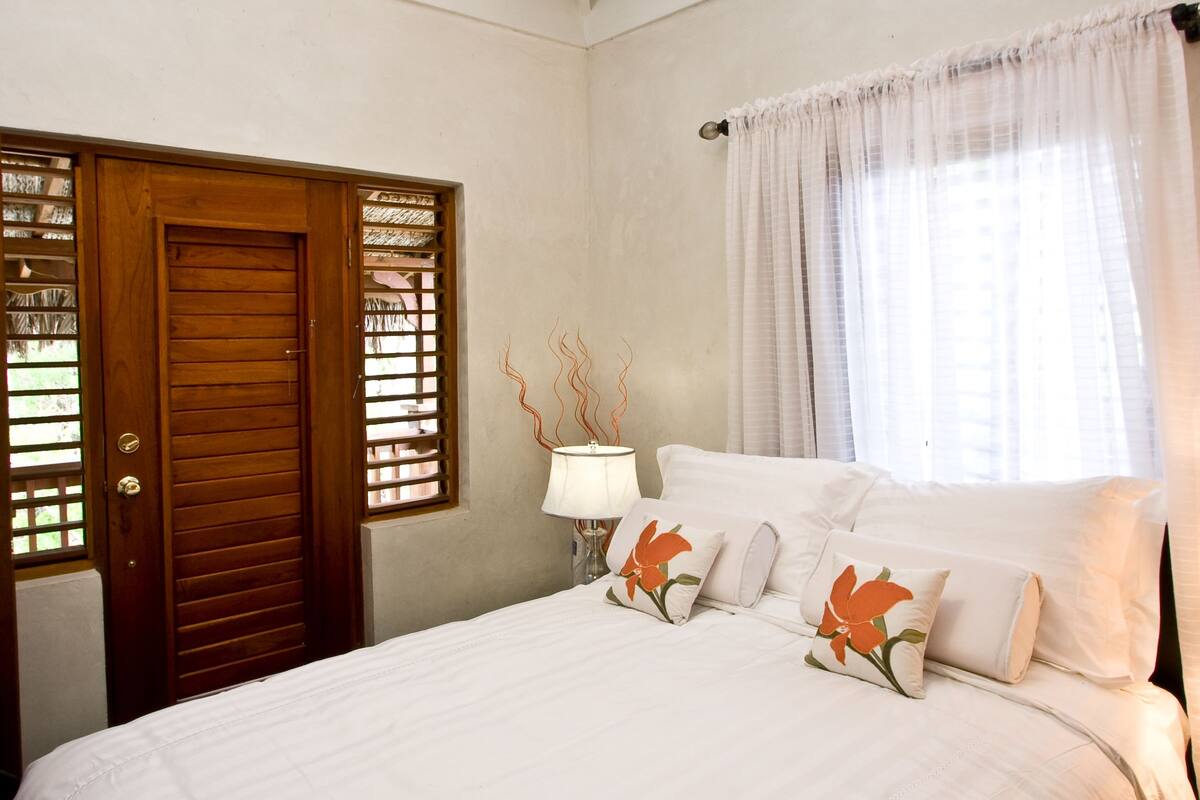 A bedroom with a large bed, a ceiling fan, a wooden door, and a window with curtains.