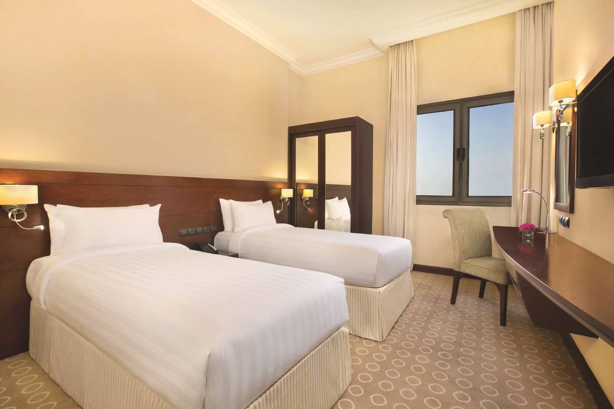Una habitación de hotel con dos camas, un escritorio, una silla, un espejo grande y una ventana.