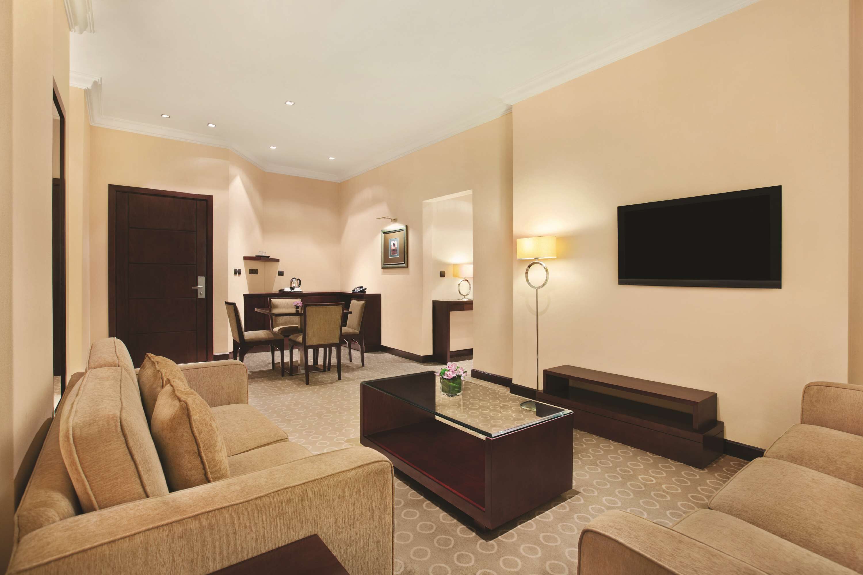 A hotel room with a sofa, a coffee table, a TV, a desk with chairs, and a lamp.