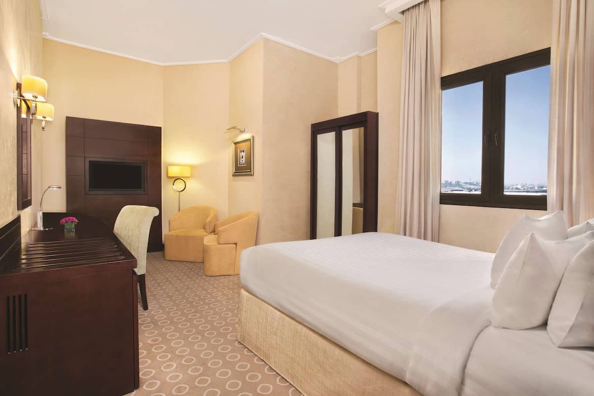 Habitación de hotel con una cama grande, un escritorio con silla, un televisor y vista a la ciudad desde la ventana.