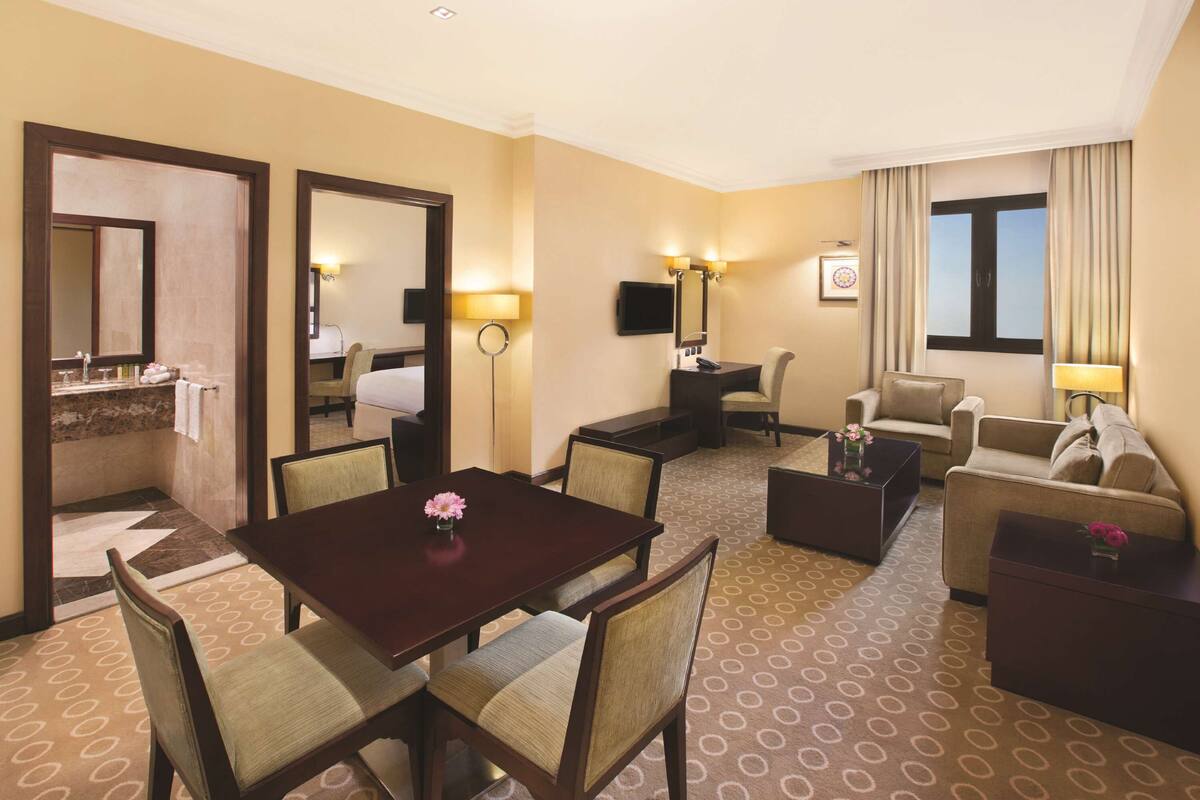 Habitación de hotel con una mesa de comedor, dos sillones, un sofá, un televisor y un ventanal con cortinas.