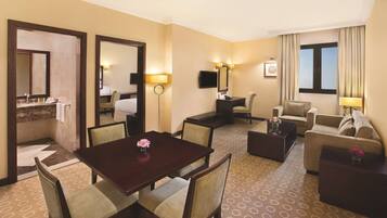 A hotel room with a dining table, two armchairs, a sofa, a TV, and a large window with curtains.