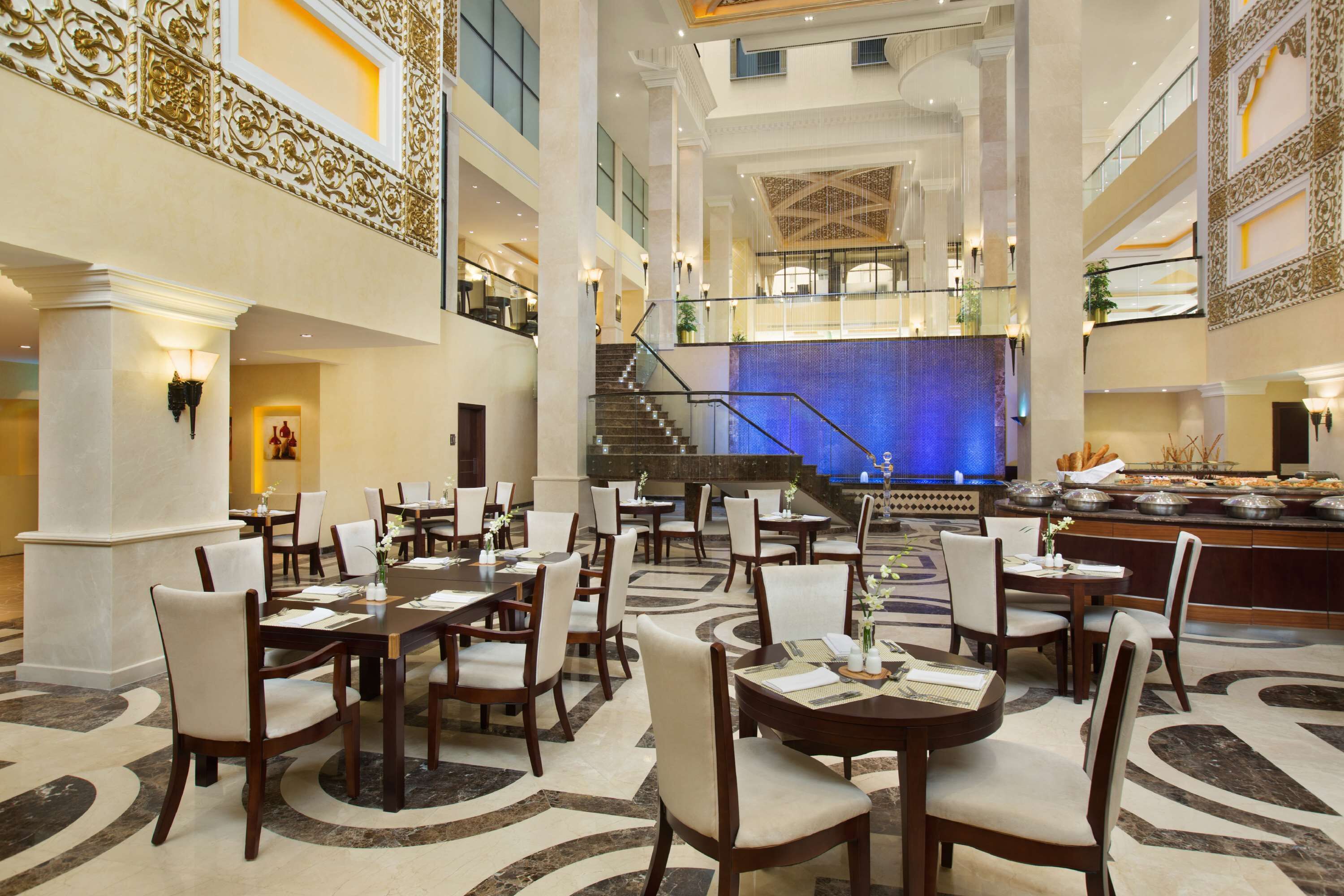 Een ruime hotellobby met meerdere eettafels en stoelen, een monumentale trap en een decoratieve fontein.