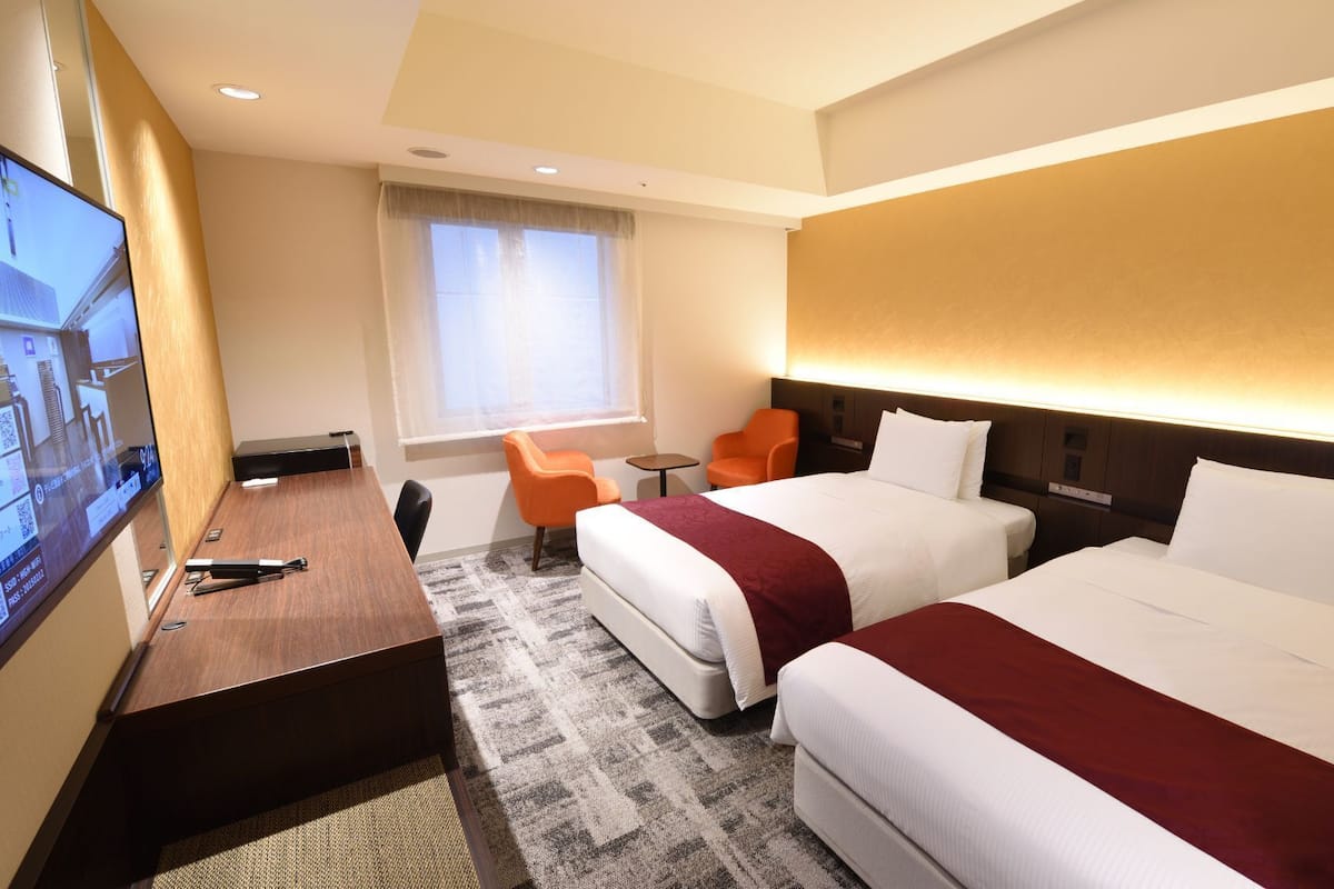 A hotel room with two beds, a desk, a TV, and a window with curtains.