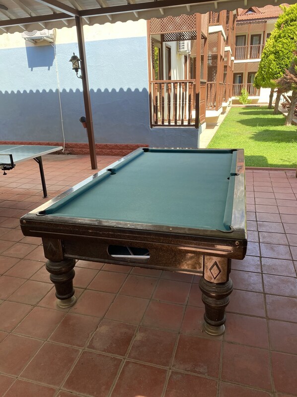 A pool table in an outdoor area with a tiled floor, a covered seating area, and a building in the background.