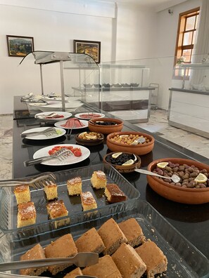 Un bufé con diversos platos y postres, entre ellos pasteles y aceitunas, dispuesto en una mesa con vitrinas de vidrio.