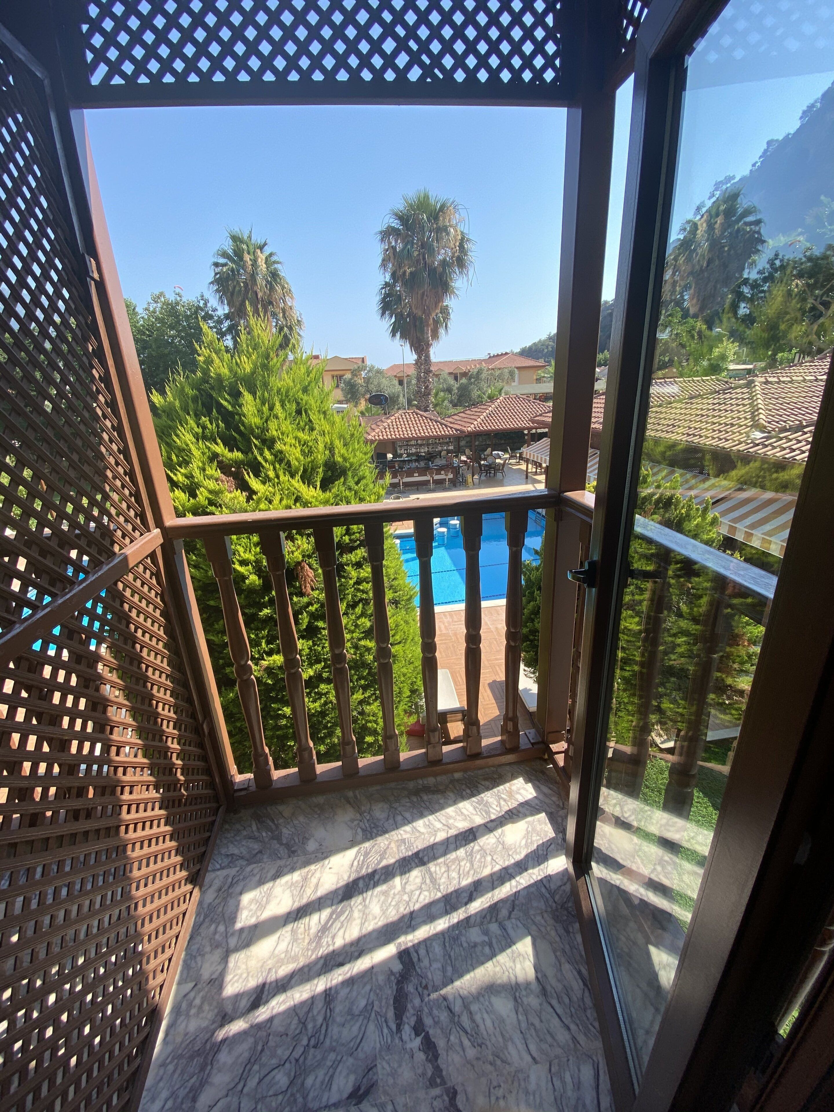 A balcony with a view of a swimming pool, surrounded by greenery and buildings with tiled roofs.