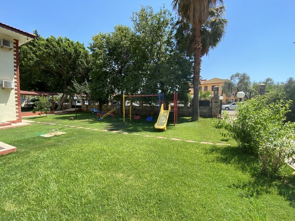 A grassy backyard with a playground, including a slide and swings, surrounded by trees and a building.