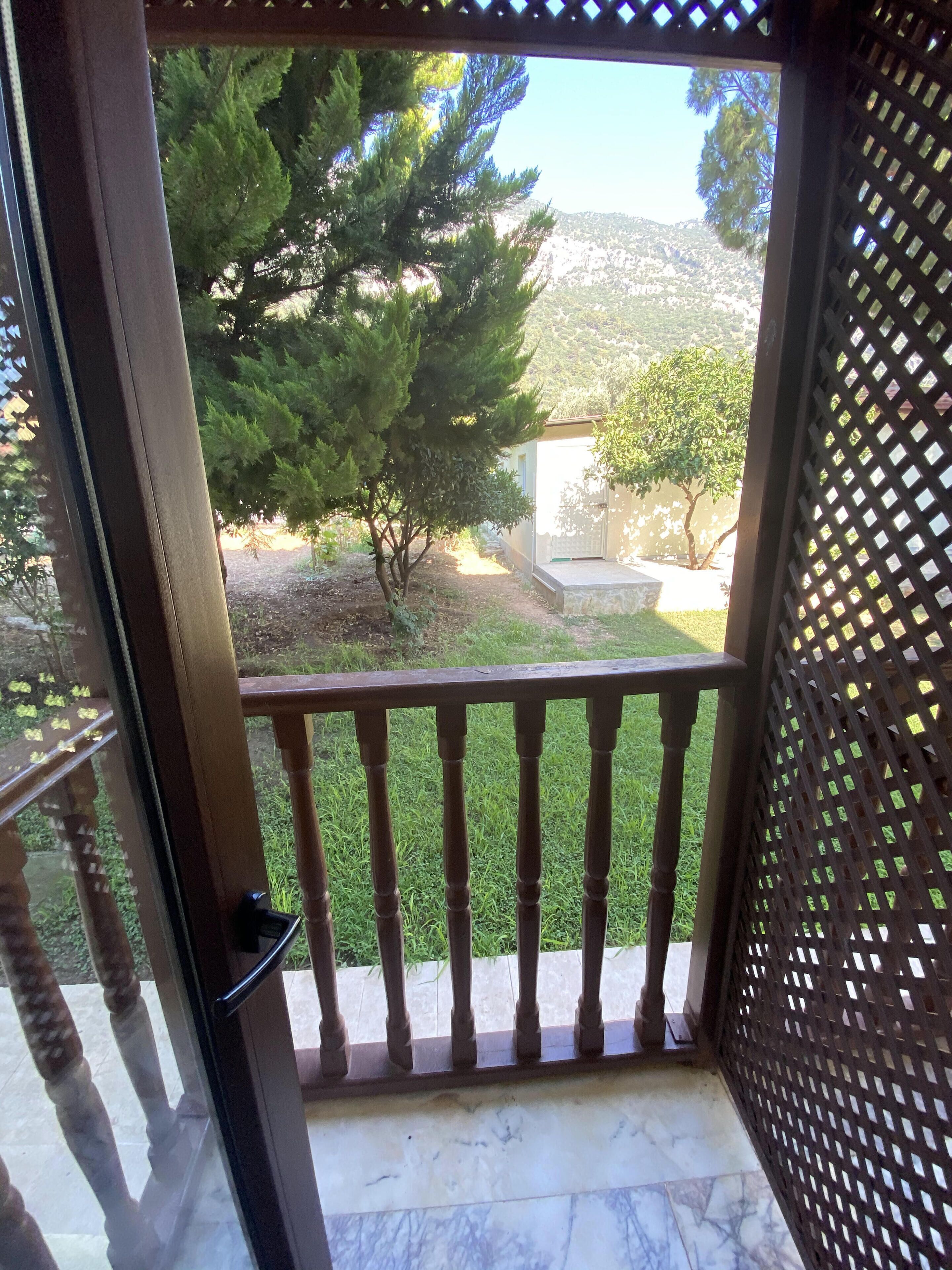 Ein aus Holz gestalteter Balkon mit Gittergeländer, einem Marmorboden und Blick auf einen Garten mit Bäumen sowie ein Gebäude im Hintergrund.