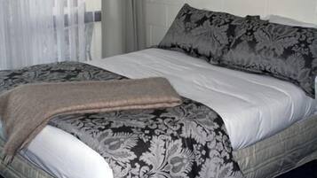 A neatly made bed with a patterned bedspread and pillows, a bedside table, and a window with sheer curtains.