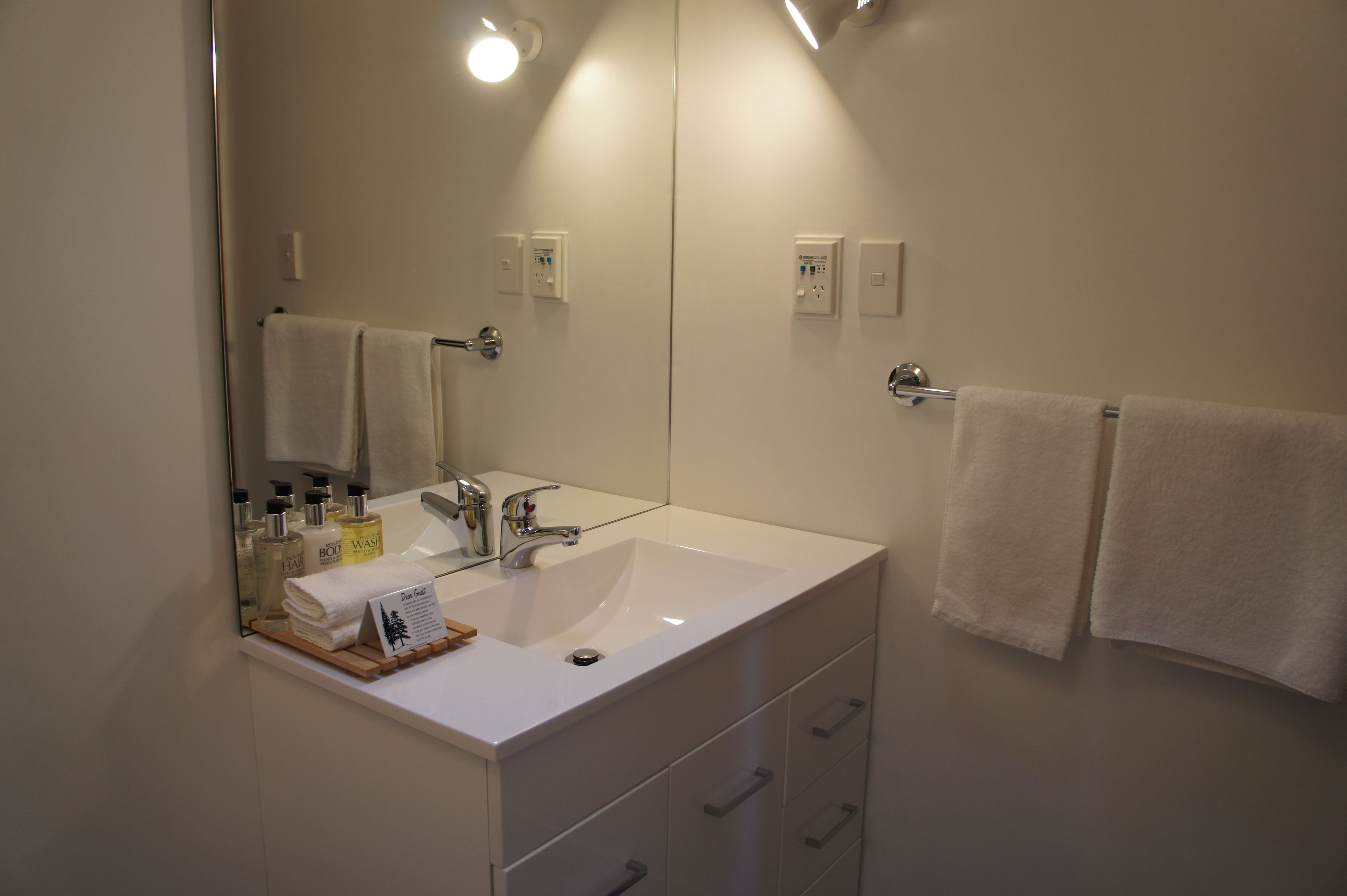 Un baño con lavamanos blanco, espejo y toallero. Hay artículos de aseo en la encimera.