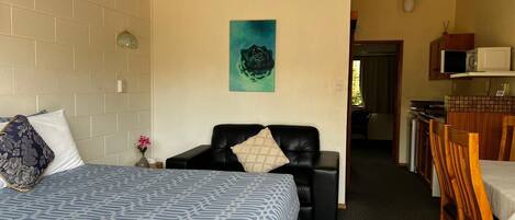 A room with a bed, a black leather chair, a dining table with chairs, a microwave, and a painting on the wall.