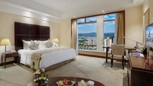 Ein Hotelzimmer mit einem großen Bett, einem Schreibtisch, einem Stuhl und Blick auf eine Stadtlandschaft durch große Fenster.