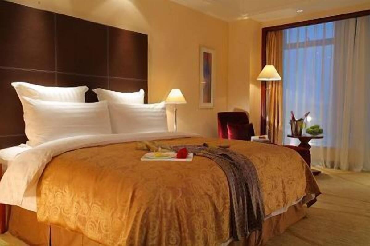 A hotel room with a large bed, a nightstand, a lamp, and a view of the outdoors through a window.