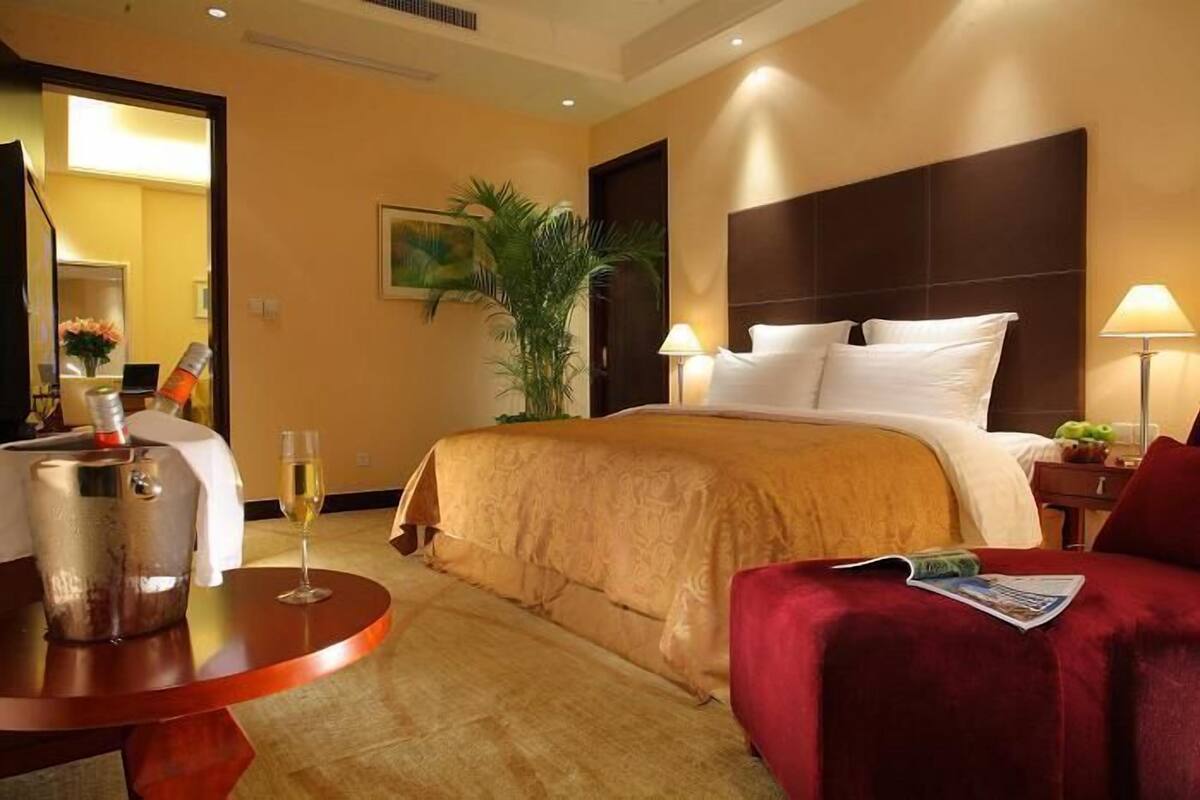 A hotel room with a large bed, a round table with a bottle and glass, a red armchair, and a nightstand with a lamp.