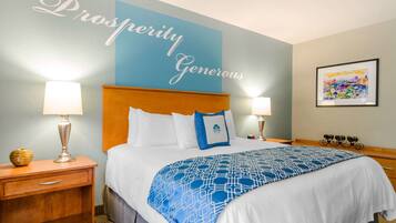 A hotel room with a large bed, two bedside tables, a nightstand, a wall with a 'Prosperity Generous' sign, and a framed artwork on the wall.