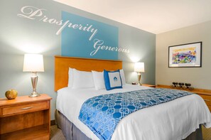 A hotel room with a large bed, two bedside tables, a nightstand, a wall with a 'Prosperity Generous' sign, and a framed artwork on the wall.