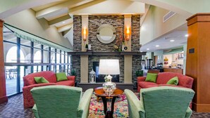 A lobby with a stone fireplace, two green armchairs, a round table, and a large window.