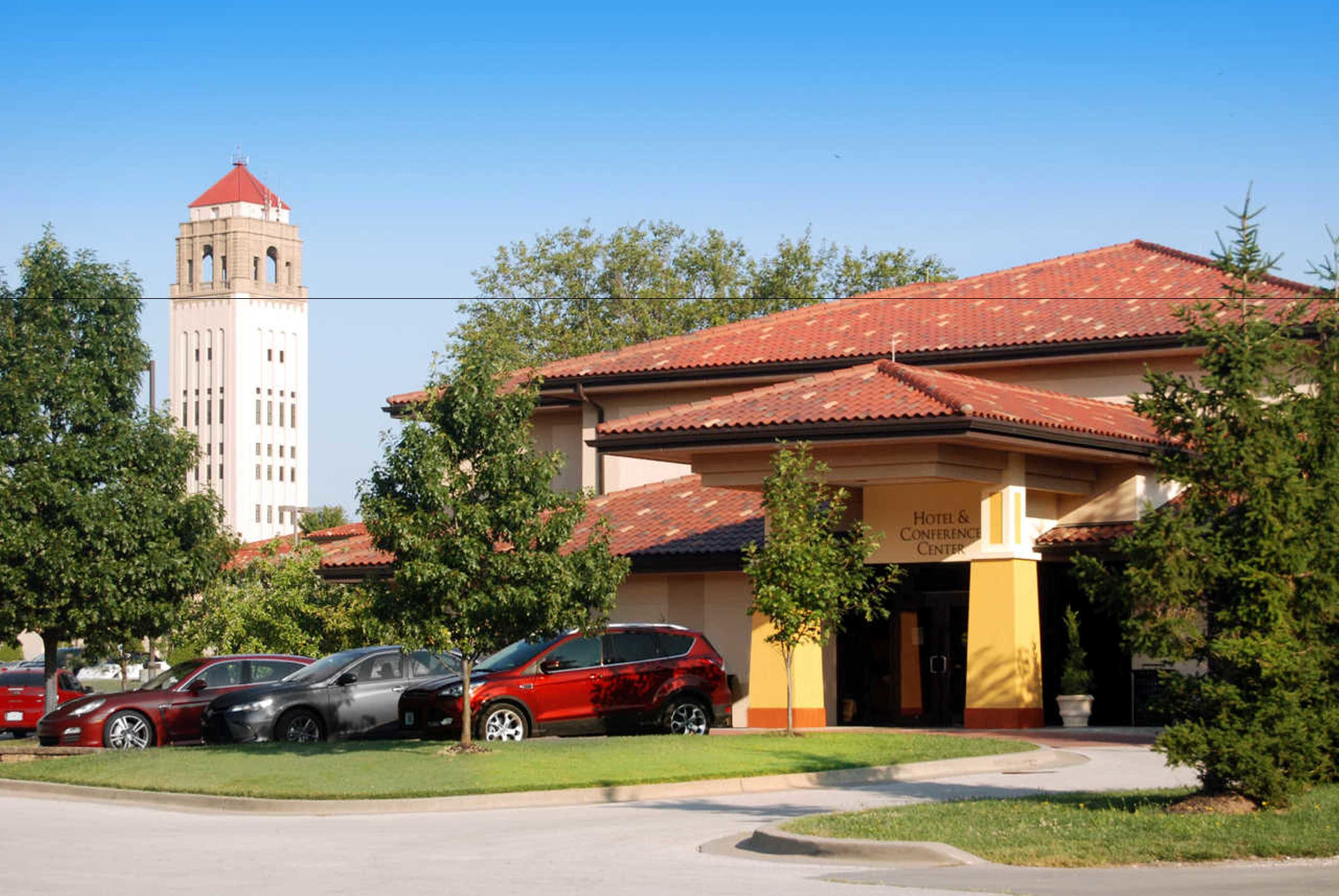 A hotel with a red-tiled roof, a tower in the background, and several parked cars.