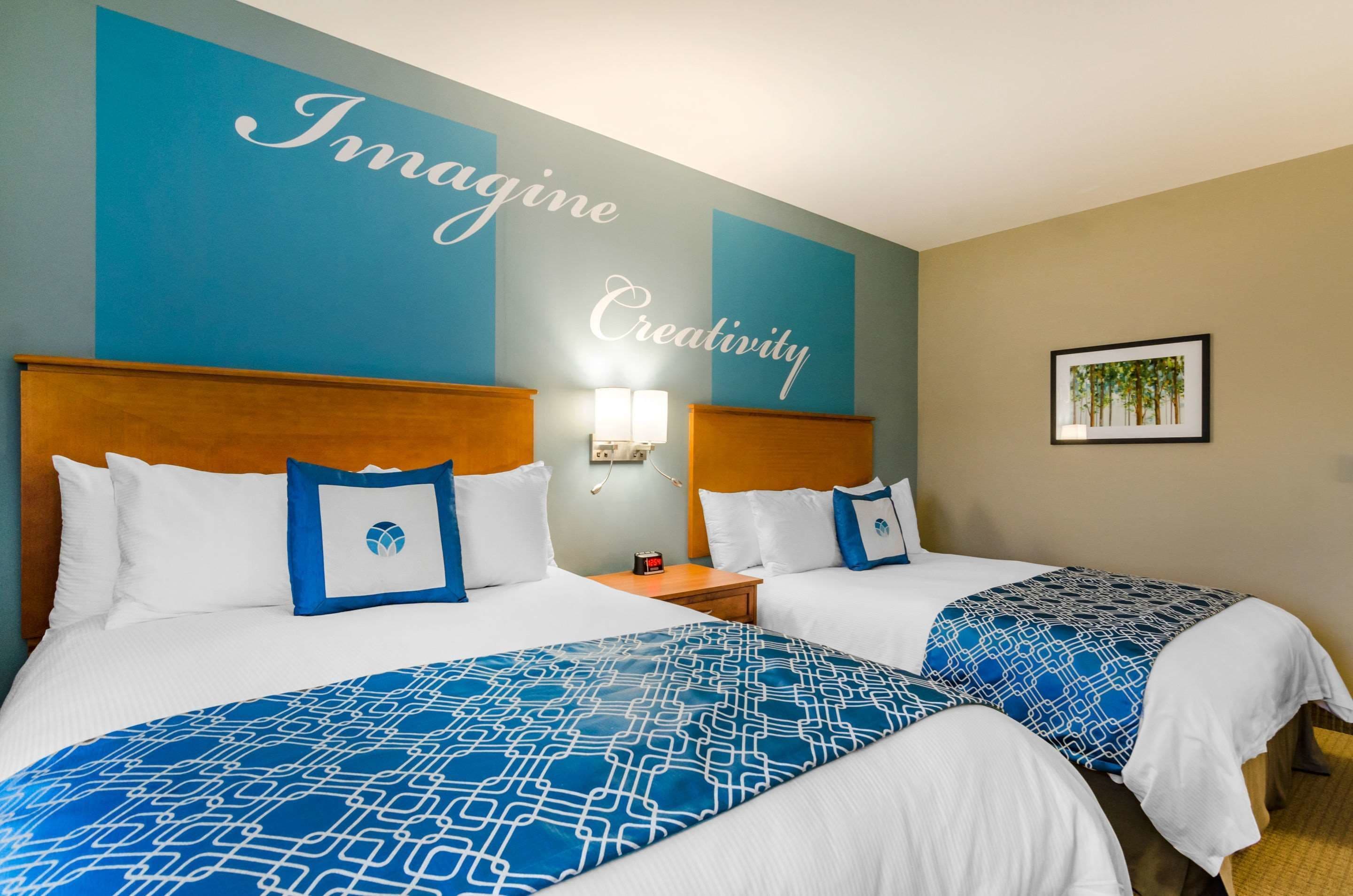 A hotel room with two beds, each with a blue patterned bedspread and white pillows. There is a wall with the words "Imagine Creativity" and a framed picture of trees on the wall.