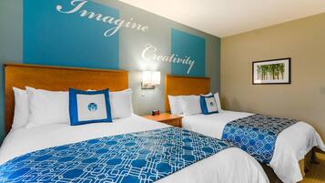 A hotel room with two beds, each with a blue patterned bedspread and white pillows. There is a wall with the words "Imagine Creativity" and a framed picture of trees on the wall.