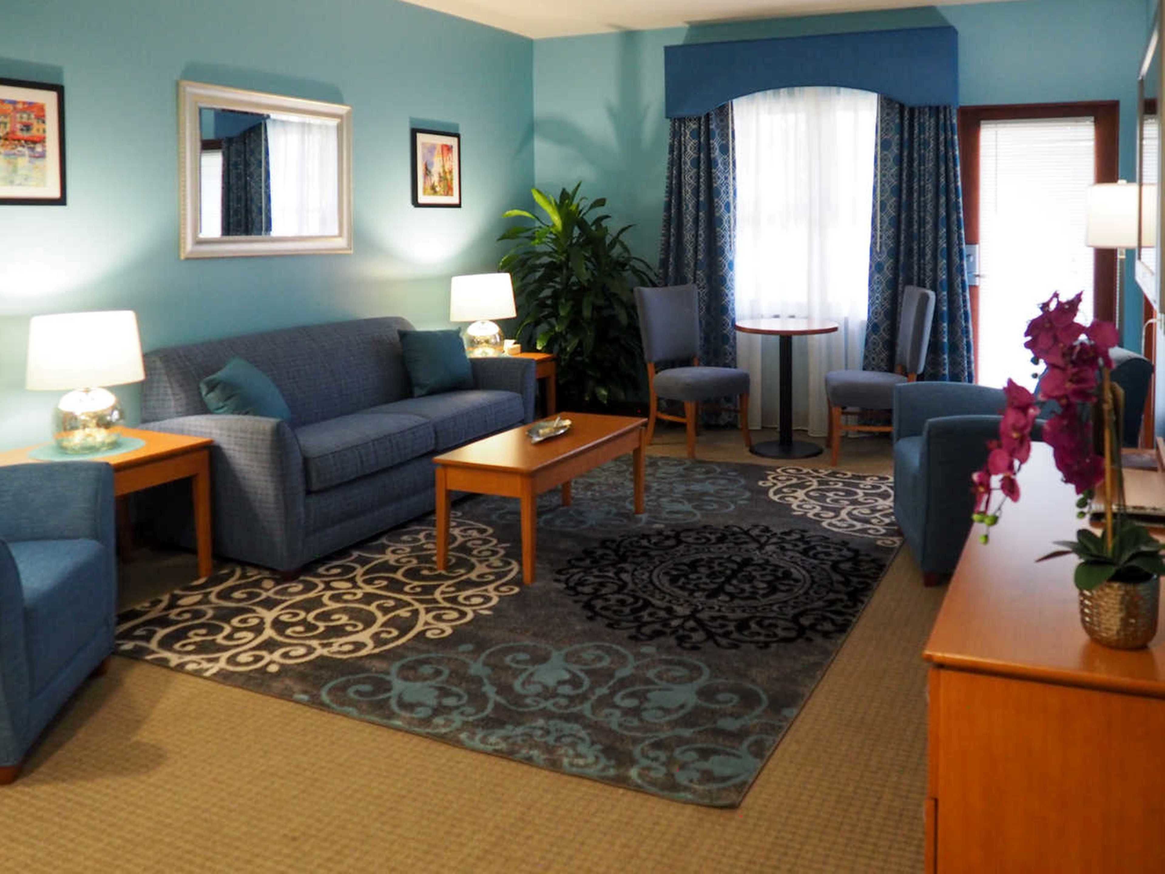 A living room with a sofa, two armchairs, a coffee table, a side table with a lamp, a potted plant, and a patterned rug.