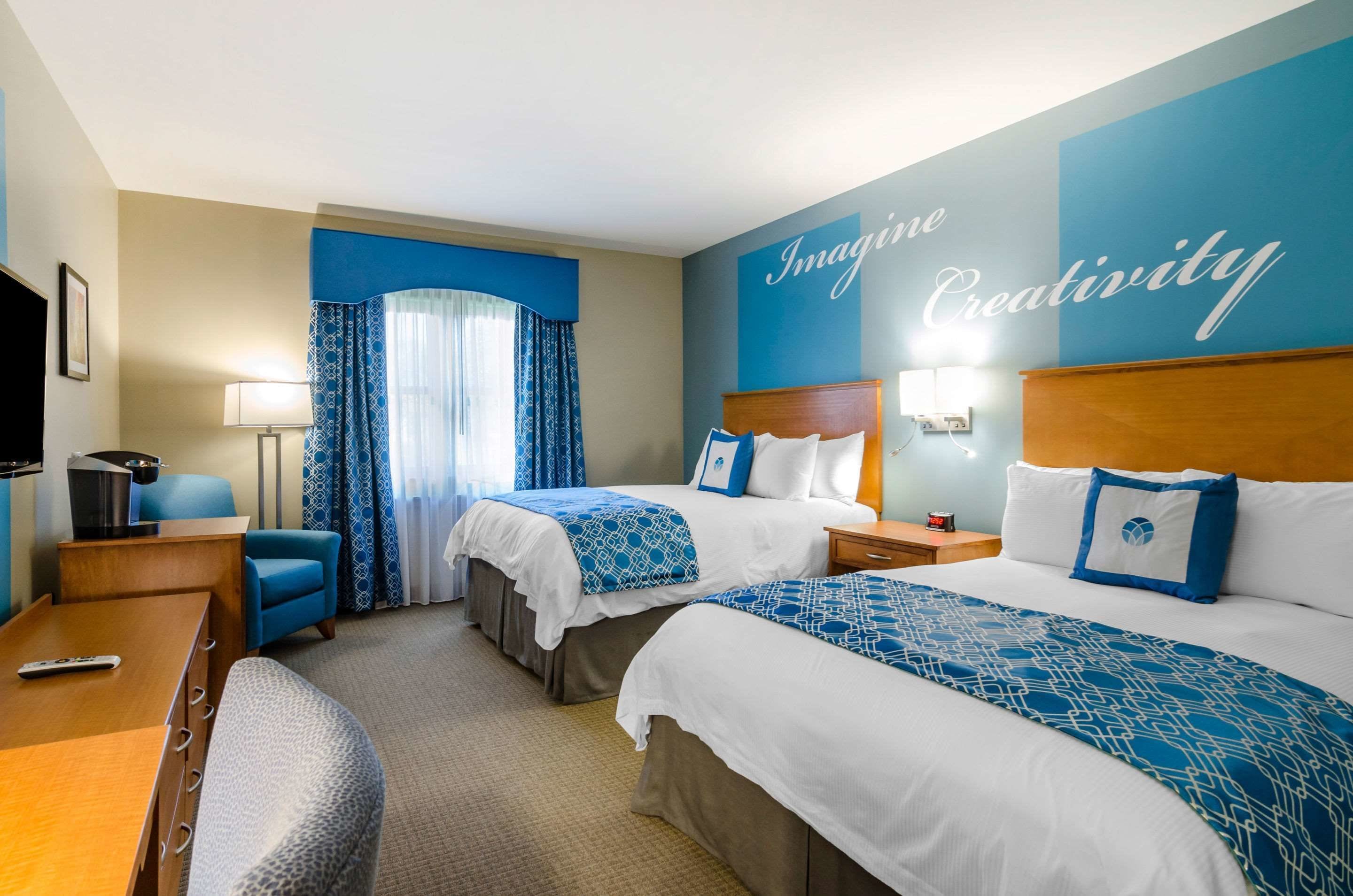 Habitación de hotel con dos camas, un escritorio, una silla, un televisor y una pared que dice 'Imagine Creativity'.