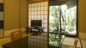 Um quarto japonês tradicional com tatames, uma televisão sobre uma mesinha baixa e vista para bambuzais lá fora.