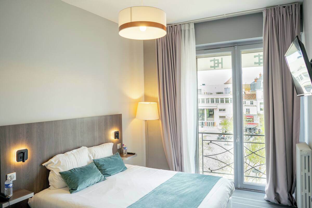 A hotel room with a bed, bedside lamps, a view of the city through a large window, and a flat-screen TV mounted on the wall.